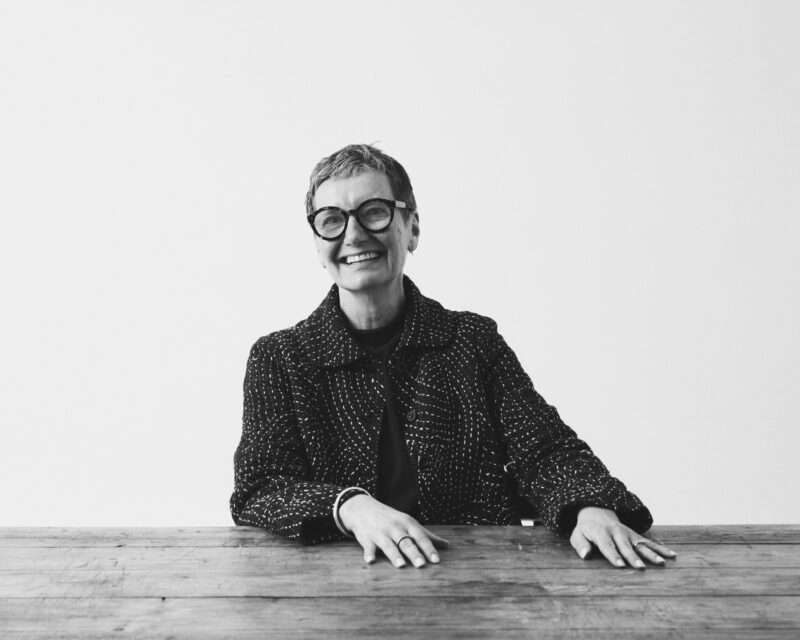 Suzie de Rohan Willner, with short hair and glasses, sits at a wooden table, smiling. The image is black and white with a plain background.