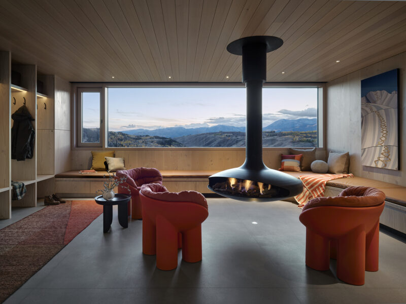 Modern living room with orange rounded chairs and a suspended fireplace facing a large window overlooking mountain hills.