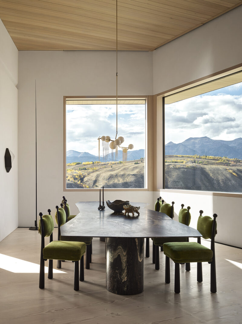 A dining room with a stone-top table and green upholstered chairs, featuring large windows with a view of mountains and a valley.
