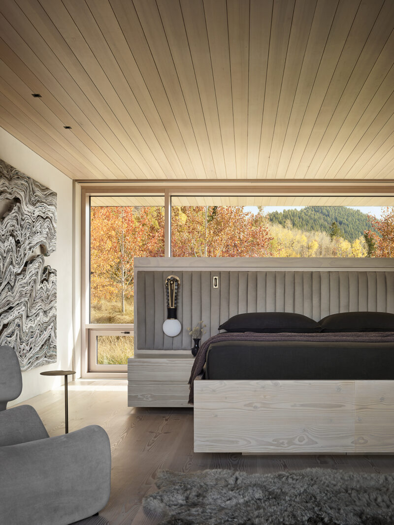 Modern bedroom with a light wood platform bed, upholstered headboard, and large window overlooking an autumn forest.